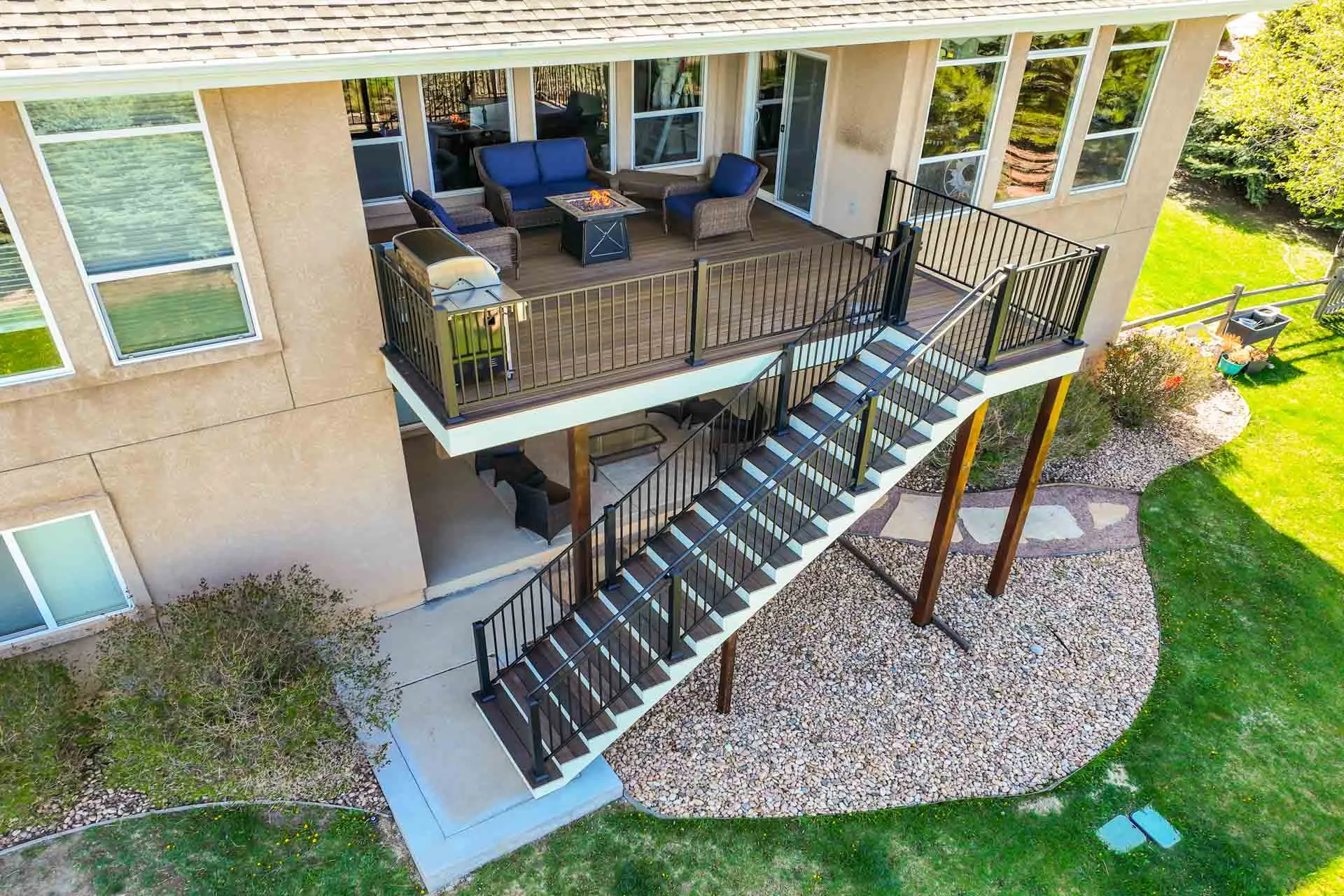 A beige house with an elevated deck and black metal railing, blue-cushioned outdoor furniture around a fire pit, connected by a staircase to a gravel yard below.