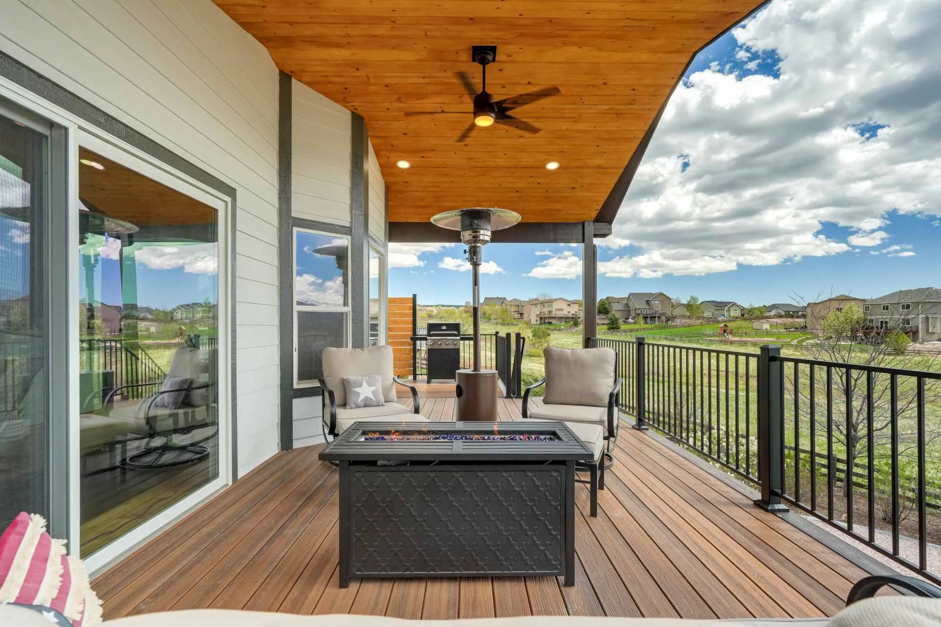 Covered wooden-deck patio with ceiling fan, outdoor heater, and two cushioned chairs facing a fire pit table and a grill, overlooking a suburban yard.