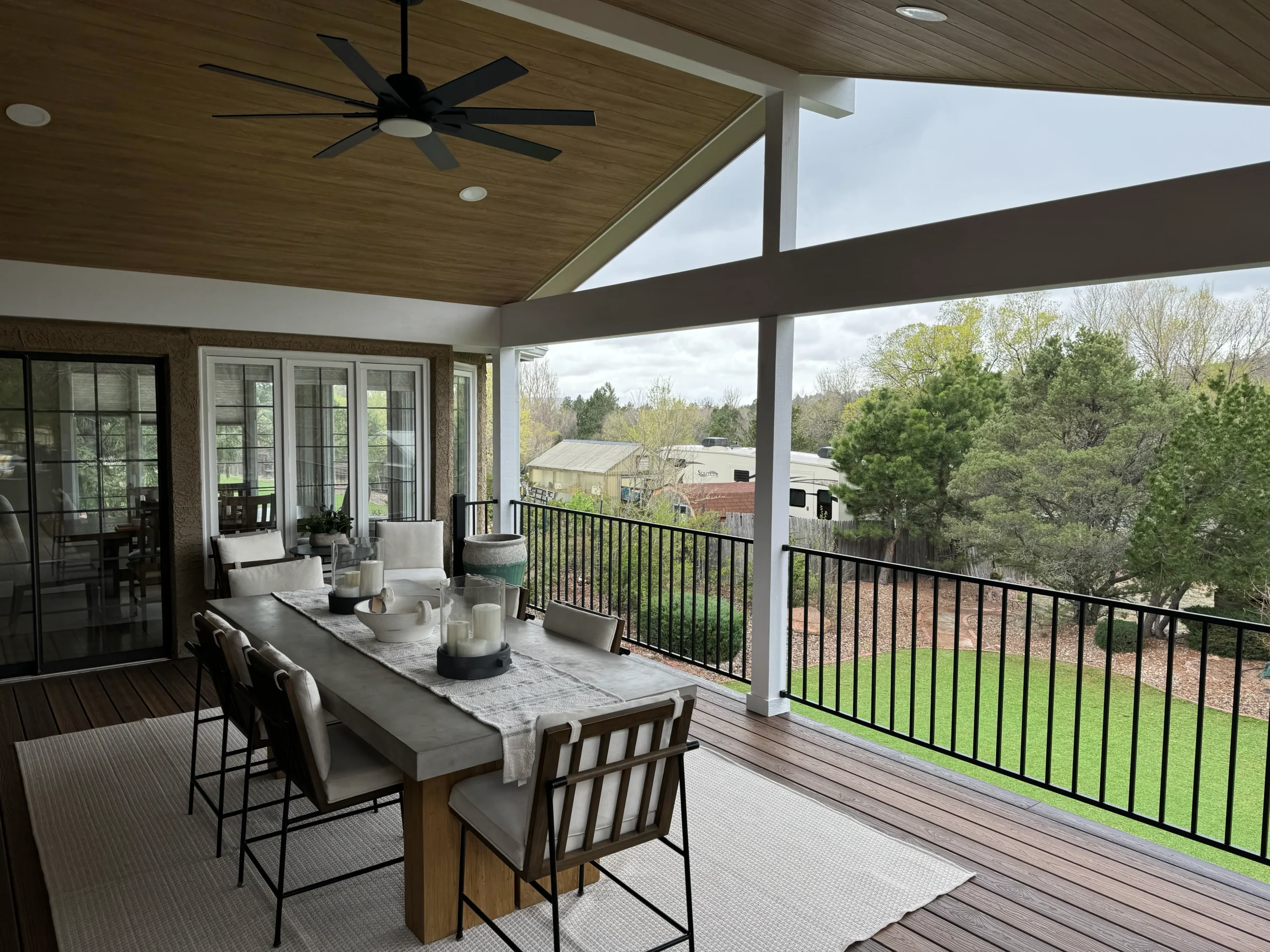 Covered patio with a wooden ceiling, black ceiling fan, and an outdoor dining table set with candles on a cream runner, overlooking a green yard and trees behind a black railing.